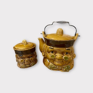 Puede incluir: Un juego vintage de tetera y azucarero de cerámica con caras caprichosas con gafas y pajaritas. La tetera tiene una tapa marrón y un asa de resorte. El azucarero tiene una tapa marrón.