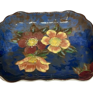 May include: Rectangular ceramic serving dish with a wavy edge. The dish is painted with a deep blue background and features three large flowers in shades of red, yellow, and orange. The flowers have green leaves and yellow centres.