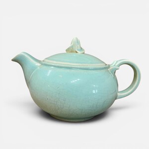 May include: A light blue ceramic teapot with a lid and handle. The teapot has a crackle glaze finish and a small decorative element on the lid. The spout is curved, and the handle is rounded.