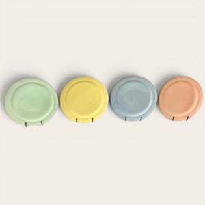 Puede incluir: Cuatro platos de cerámica coloridos en tonos pastel de verde, amarillo, azul y melocotón. Cada plato tiene un diseño circular con un centro elevado y un pequeño gancho debajo. Los platos están dispuestos en fila sobre un fondo neutro.