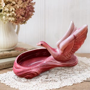 May include: A glossy, deep rose-pink ceramic swan planter featuring a sculpted bird with raised wings and a smooth, curved basin. The swan has a dark eye detail and a marbled, multi-tonal pink glaze finish that highlights the molded feather textures.