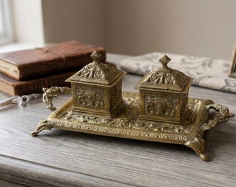 May include: A rectangular brass tray with ornate scrollwork handles and feet, featuring two matching square lidded containers. Each container has a pointed finial and detailed relief carvings on all sides, creating a cohesive metal desk set.
