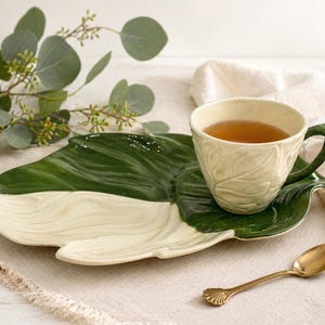 May include: A ceramic teacup and saucer set with a leaf design. The teacup is filled with tea and sits on a green and cream-coloured leaf-shaped saucer. A gold spoon rests beside the saucer. The set is on a white surface.
