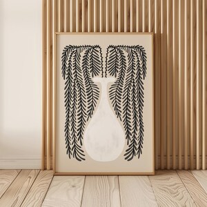 May include: A framed art print featuring a minimalist illustration of a white vase with black weeping willow branches. The artwork is set against a beige background and framed in a light brown wooden frame. The print is displayed on a wooden floor.