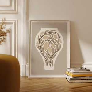 May include: Framed art print featuring a beige vase design with brown leaves and tan fruit illustrations. The artwork is set against a light gray background and framed in white. The print is leaning against a white wall and wooden floor.