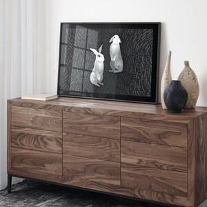 May include: A framed rectangular print featuring two white rabbits against a dark, textured background with small white dots. The print is displayed on a wooden dresser with nine drawers, accompanied by three decorative vases of varying shapes.