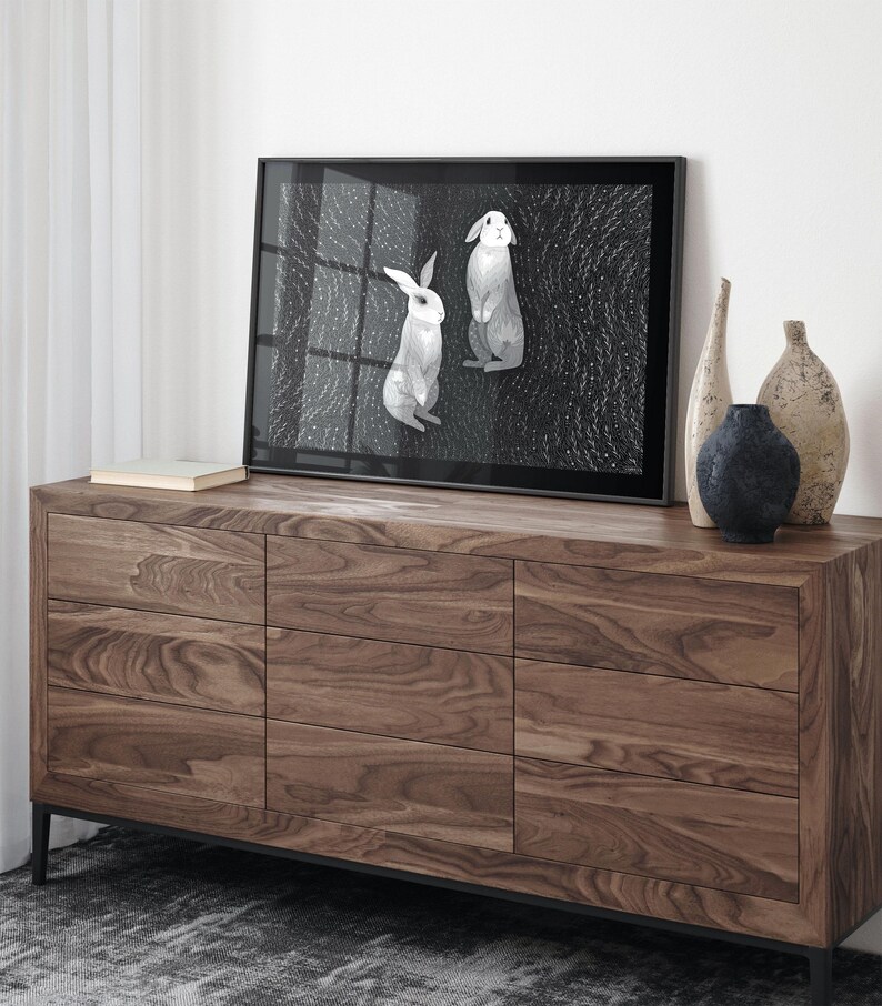 May include: A framed rectangular print featuring two white rabbits against a dark, textured background with small white dots. The print is displayed on a wooden dresser with nine drawers, accompanied by three decorative vases of varying shapes.