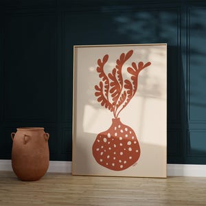 May include: A framed art print featuring a terracotta-colored abstract design of a vase with a plant. The vase has white polka dots. The artwork is displayed in a room with dark teal walls and a wooden floor.