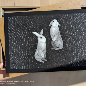 May include: A rectangular art print featuring two grey and white rabbits centered against a dark background filled with intricate white line patterns of swirling foliage and small dots. The print is set within a black mat board border.
