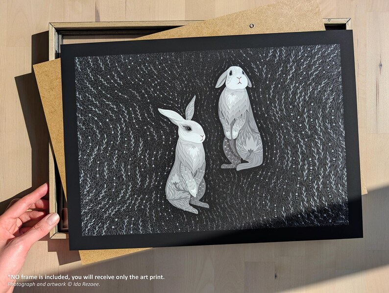 May include: A rectangular art print featuring two grey and white rabbits centered against a dark background filled with intricate white line patterns of swirling foliage and small dots. The print is set within a black mat board border.