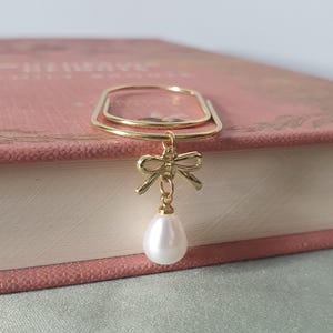 May include: A gold-coloured bookmark featuring a bow and a dangling, teardrop-shaped pearl. The bookmark is rectangular with rounded corners. The pearl is white and has a smooth surface. The bookmark rests on a pink book.