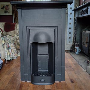 May include: A matte black cast iron fireplace surround featuring a rectangular mantelpiece, a central arched opening, and a lower grate section. The structure has clean, straight lines and a solid, uniform dark finish throughout its entire frame.