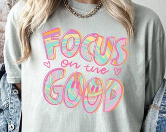 May include: A light blue t-shirt with a colorful graphic that says "Focus on the good" in a retro font.