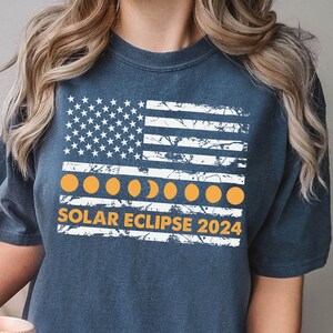 May include: Navy blue t-shirt featuring a distressed American flag design with orange eclipse phases and the text "SOLAR ECLIPSE 2024". The shirt has a vintage look and is made of soft material.