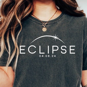 May include: A dark gray t-shirt with a white graphic of a crescent moon and a star with the text "ECLIPSE 04.08.24".