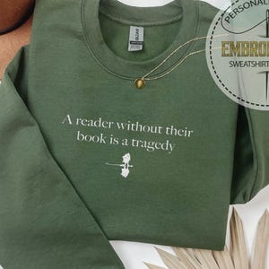 May include: An olive green sweatshirt with the quote "A reader without their book is a tragedy" in white. A small graphic of a book and feather is below the quote. A gold necklace and a brown hat are also visible.