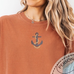 May include: Rust-colored t-shirt with an embroidered anchor design. The anchor is dark gray with gold rope detailing. The shirt has rolled-up sleeves and is paired with a denim jacket. The text "PERSONALIZED GIFT EMBROIDERED COMFORT COLORS" is visible.