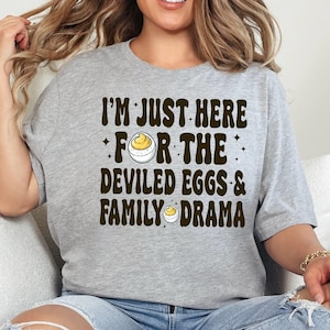 May include: A woman wearing a grey t-shirt with the text "I'm just here for the deviled eggs & family drama" in brown lettering. The shirt features a small illustration of a deviled egg.