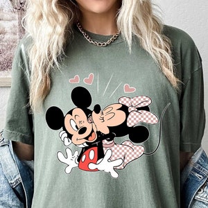 May include: A green t-shirt featuring a graphic of Mickey Mouse and Minnie Mouse kissing. The graphic is in a cartoon style and features red, black and white colours. There are pink hearts above the characters.