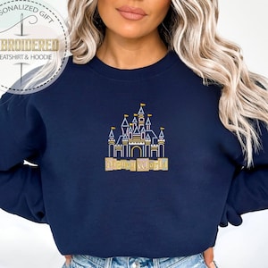 May include: Navy blue sweatshirt with an embroidered gold and white castle design and the words "Disney World". The sweatshirt has a crew neck and long sleeves. The image also includes a logo that says "Personalized Gift Embroidered Sweatshirt & Hoodie".
