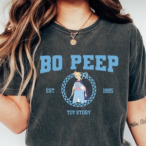 Puede incluir: Una camiseta gris oscuro con un gr&aacute;fico azul y blanco de Bo Peep de Toy Story. El gr&aacute;fico incluye el texto "Bo Peep", "Est. 1995", y "Toy Story".