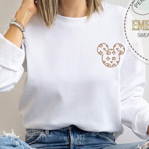 May include: White sweatshirt with a brown Mickey Mouse design on the chest. The design is made up of various shapes and patterns. The sweatshirt has long sleeves and a round neckline. The image also includes a circular graphic that says "Personalized Gift" and "Embroidered Sweatshirt & Hoodie".