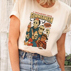 May include: Cream-colored t-shirt with a graphic that reads "I Freaking Love Summerween." The graphic features cartoon versions of horror movie characters holding ice cream cones. The shirt is paired with blue jeans.