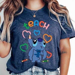 May include: A navy blue t-shirt featuring the word "teach" in rainbow colours and a blue cartoon character holding a pencil. The character is surrounded by colourful hearts and crayons.