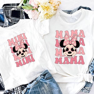 K&ouml;nnte beinhalten: Zwei wei&szlig;e T-Shirts mit einem rosa-wei&szlig;en Minnie Mouse-Design. Das gr&ouml;&szlig;ere Shirt sagt "MAMA" und das kleinere Shirt sagt "MINI".