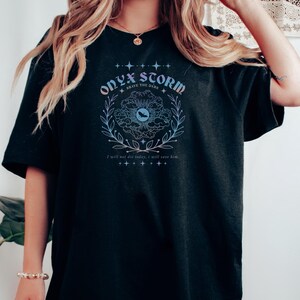 Peut inclure: T-shirt noir surdimensionn&eacute; avec un imprim&eacute; graphique d'un &oelig;il stylis&eacute; avec une lune croissante et le texte "ONYX STORM BRAVE THE DARK" dans une police gothique. Le graphique est entour&eacute; d'une couronne de feuilles et d'&eacute;toiles.