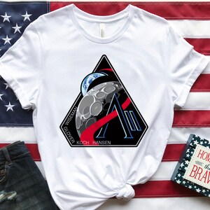 May include: White t-shirt featuring a black, red, and blue graphic of a moon, Earth, and the letter "A". The shirt is displayed on an American flag. A pair of denim shorts and a small book with the text "HOME OF THE BRAVE" are also present.