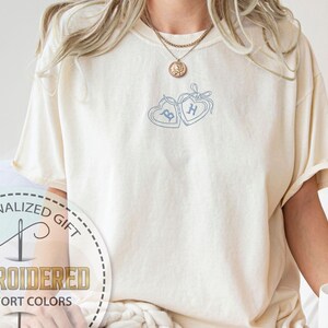 May include: Cream-colored t-shirt with embroidered blue heart design and initials. The shirt features the text "Embroidered Comfort Colors" and "Personalized Gift" in a circular logo. A person is holding a white mug.