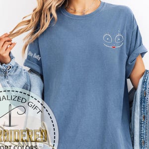 K&ouml;nnte beinhalten: Ein blaues Comfort-Colors-T-Shirt mit einem gestickten Gesichtsdesign auf der Brust. Das Shirt hat hochgekrempelte &Auml;rmel und den Text "His Sally" auf dem &Auml;rmel gestickt. Das Shirt ist Teil einer personalisierten Geschenk-Kollektion.