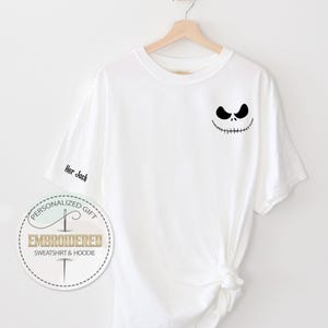 K&ouml;nnte beinhalten: Wei&szlig;es T-Shirt mit einem schwarzen Jack Skellington-Gesicht auf der Brust. Auf dem linken &Auml;rmel steht der gestickte Text "Her Jack". Das Shirt h&auml;ngt an einem Holzb&uuml;gel vor wei&szlig;em Hintergrund. Ein kreisf&ouml;rmiges Logo ist sichtbar.