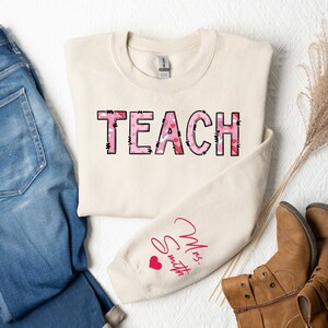 May include: A beige sweatshirt with the word "TEACH" printed in pink and black on the front. The word is made up of pink hearts with black outlines. The sleeve has the text "Mrs. Smith" printed in pink with a small pink heart.