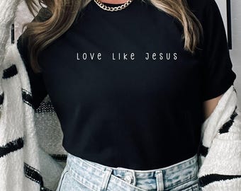 May include: A woman wearing a black t-shirt with the text "Love Like Jesus" in white lettering. She is also wearing a white and black striped cardigan and blue jeans.