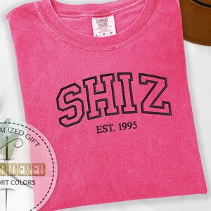 May include: A bright pink Comfort Colors t-shirt with the word "SHIZ" in black block letters and "EST. 1995" below. The shirt is folded and has a vintage look. A brown hat is partially visible in the background.