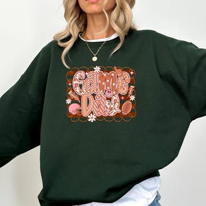 May include: Dark green sweatshirt with the words "Game Day" in a retro design. The graphic includes a football, flowers, and a skull. The sweatshirt is a comfortable, casual style.