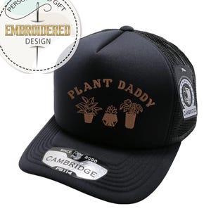 May include: Black trucker hat with the words "PLANT DADDY" in brown lettering, along with three plant illustrations. The hat has a mesh back and a curved brim. A sticker on the brim reads "CAMBRIDGE" and "PB114".