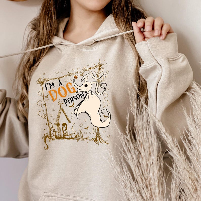 May include: Beige hoodie with a drawstring. The front features a graphic of a ghost dog with the text "I'M A DOG PERSON" in orange and black. The design includes a house and decorative elements in brown.
