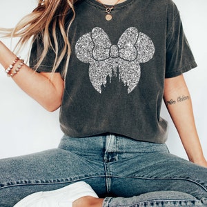 Puede incluir: Una camiseta gris oscuro con un lazo de purpurina plateada y una silueta de castillo dentro del lazo. La camiseta la lleva puesta una persona que lleva pantalones vaqueros azules y zapatillas blancas.