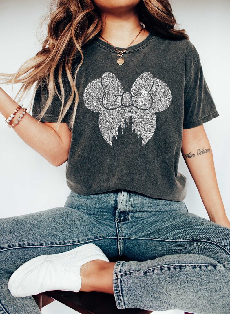Puede incluir: Una camiseta gris oscuro con un lazo de purpurina plateada y una silueta de castillo dentro del lazo. La camiseta la lleva puesta una persona que lleva pantalones vaqueros azules y zapatillas blancas.