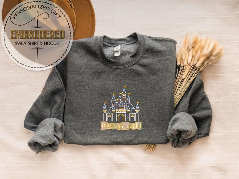 May include: A dark gray sweatshirt with an embroidered castle design and the words "Disney World" in gold. The sweatshirt has long sleeves and a crew neck. The image also includes text that says "Personalized Gift" and "Embroidered Sweatshirt & Hoodie".