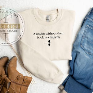 May include: Cream-colored sweatshirt with the text "A reader without their book is a tragedy" in black. The image includes blue jeans and brown leather boots, suggesting a casual, everyday style. The sweatshirt is a personalized gift.
