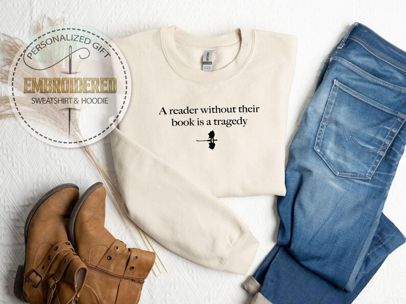 May include: Cream-colored sweatshirt with the text "A reader without their book is a tragedy" in black. The image includes blue jeans and brown leather boots, suggesting a casual, everyday style. The sweatshirt is a personalized gift.