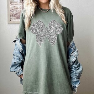 Puede incluir: Una camiseta verde de gran tama&ntilde;o con un dise&ntilde;o de lazo de purpurina plateada.