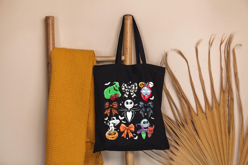 May include: Black tote bag featuring colorful illustrations of characters from The Nightmare Before Christmas. The design includes Jack Skellington, Sally, Oogie Boogie, and other iconic elements. The bag is hanging on a wooden ladder.
