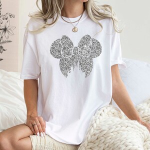 Puede incluir: Una camiseta blanca con un dise&ntilde;o de lazo negro hecho de peque&ntilde;os puntos. El lazo est&aacute; centrado en la camiseta y es el elemento principal del dise&ntilde;o.