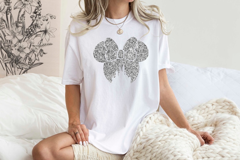 Puede incluir: Una camiseta blanca con un dise&ntilde;o de lazo negro hecho de peque&ntilde;os puntos. El lazo est&aacute; centrado en la camiseta y es el elemento principal del dise&ntilde;o.