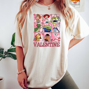 K&ouml;nnte beinhalten: Ein wei&szlig;es T-Shirt mit einem Aufdruck von Toy Story-Charakteren in rosafarbenen Herzen. Der Text "My Valentine" befindet sich unter den Figuren.